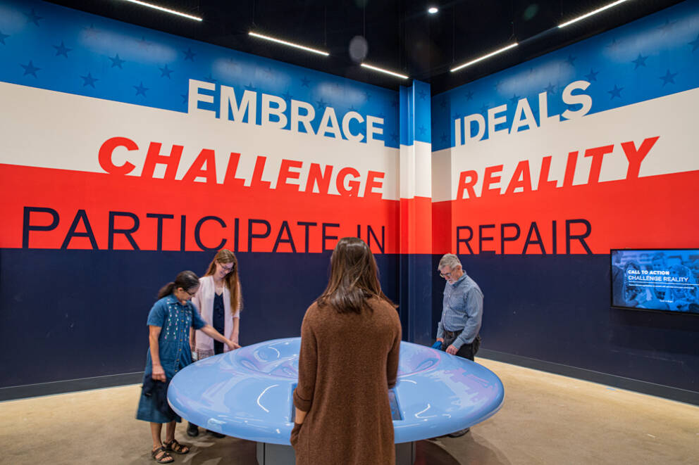 four people stand in front of touchscreens at a circular table. On the wall behind is painted in large letters, 